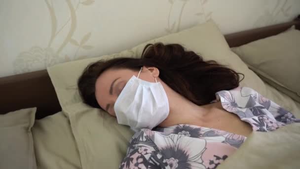 Alone Caucasian woman in floral night blouse wear blue protection medical face mask on the bed at home. Sick girl with high temperature sweetly sleeps and recover her health on a sunny day close up. - Footage, Video