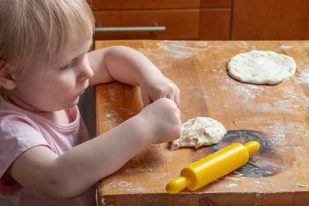 Little baby girl kneads the dough on a wooden table with hands and a rolling pin. The child is happy that he can cook pastries on his own. Baby's face and clothes in flour. Happy childhood. - Photo, image