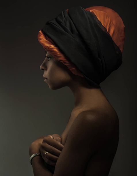 Африканская женщина стоит на черном фоне, держа грудь руками и отводя взгляд, с национальным шарфом на голове
 - Фото, изображение
