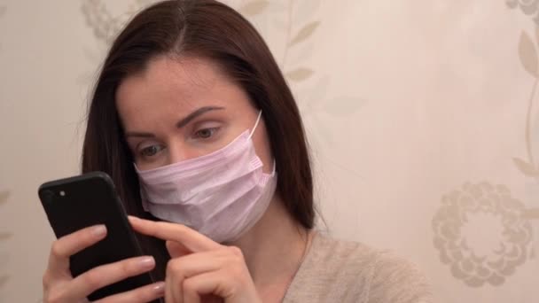 Beautiful young woman in protective pink face mask relaxing at home and using mobile black phone close up. Online chat. Social networks. Quarantine during isolation period from Covid-19 virus threat. - Záběry, video
