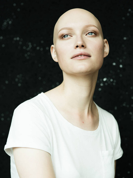 A young girl without hair in a white t-shirt, on a black background, looks away with a smile - Photo, Image
