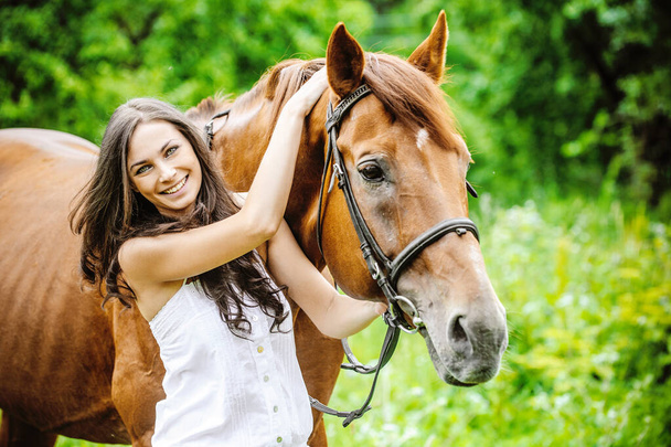 Женщина в белом платье держит коричневую лошадь на фоне летнего зеленого парка
. - Фото, изображение
