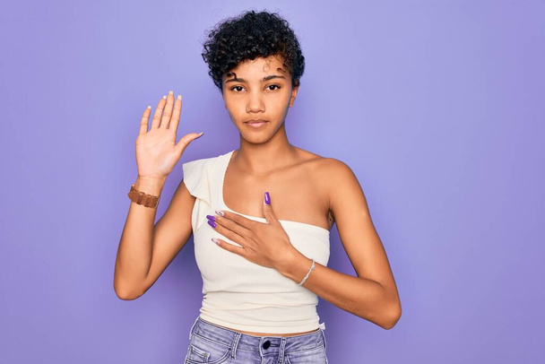 Junge schöne afrikanisch-amerikanische Afro-Frau trägt lässiges T-Shirt über lila Hintergrund Vereidigung mit der Hand auf der Brust und offener Handfläche und leistet einen Treueschwur - Foto, Bild