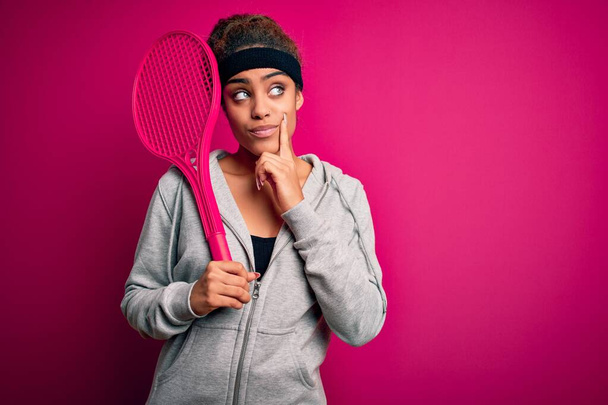 Młoda afrykańska sportowiec gra w tenisa za pomocą rakiety na odizolowanym różowym tle poważna twarz myśli o pytaniu, bardzo mylić pomysł - Zdjęcie, obraz