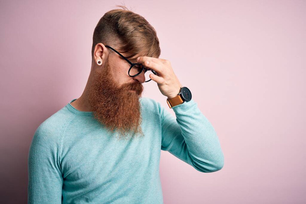 Knappe Ierse roodharige man met baard draagt bril over roze geïsoleerde achtergrond moe wrijven neus en ogen gevoel vermoeidheid en hoofdpijn. Stress en frustratie concept. - Foto, afbeelding