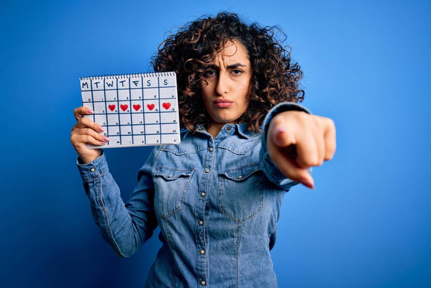 Junge schöne, lockige arabische Frau mit einem Kalender, der den Menstruationszyklus kontrolliert und mit dem Finger auf die Kamera und Sie zeigt, Handzeichen, positive und selbstbewusste Geste von vorne - Foto, Bild