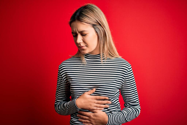 Young beautiful blonde woman wearing casual striped sweater over red isolated background with hand on stomach because indigestion, painful illness feeling unwell. Ache concept. - Foto, Bild
