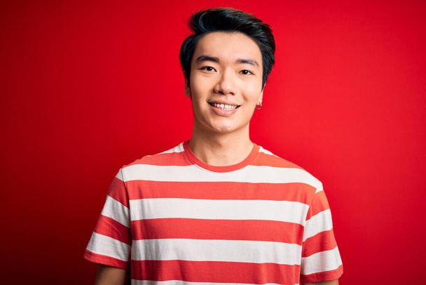 Νεαρός όμορφος Κινέζος που φοράει casual ριγέ t-shirt πάνω από κόκκινο φόντο με χαρούμενο και δροσερό χαμόγελο στο πρόσωπο. Τυχερός άνθρωπος. - Φωτογραφία, εικόνα