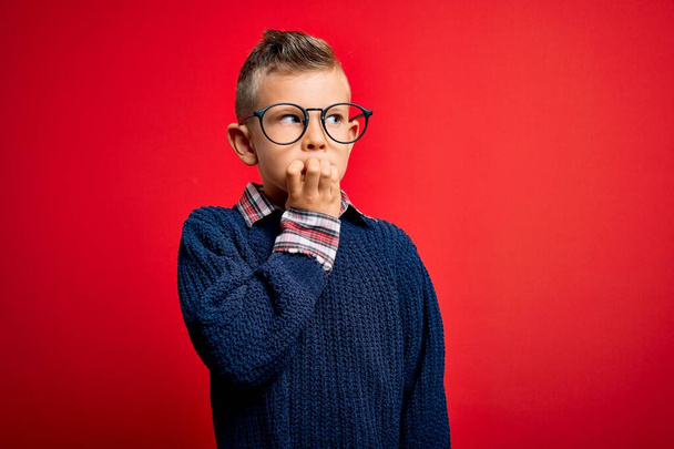 Junge kleine kaukasische Kinder mit blauen Augen stehen mit smarter Brille vor rotem Hintergrund und sehen gestresst und nervös aus, die Hände auf mundgebissenen Nägeln. Angstproblem. - Foto, Bild