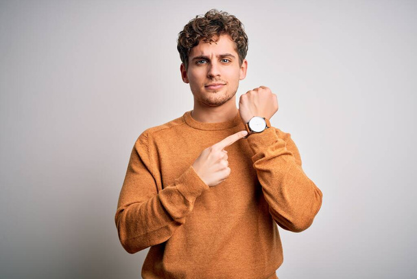 Young blond handsome man with curly hair wearing casual sweater over white background In hurry pointing to watch time, impatience, looking at the camera with relaxed expression - Foto, immagini