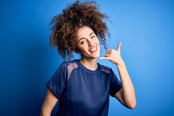 Mladá krásná žena s kudrnatými vlasy a piercing na sobě ležérní modré tričko s úsměvem dělat telefonní gesto s rukou a prsty jako mluvit do telefonu. Komunikační koncepce. - Fotografie, Obrázek