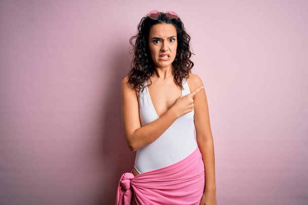 Beautiful woman with curly hair on vacation wearing white swimsuit over pink background Pointing aside worried and nervous with forefinger, concerned and surprised expression - Photo, Image