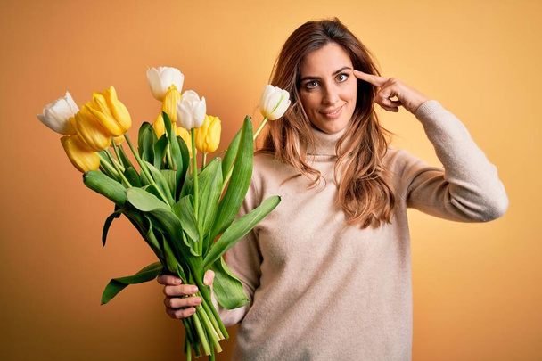 Giovane bella donna bruna in possesso di bouquet di tulipani gialli su sfondo isolato Sorridente punta alla testa con un dito, grande idea o pensiero, buona memoria
 - Foto, immagini