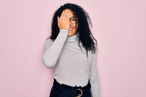 Jeune femme afro-américaine portant un pull col roulé et des lunettes sur fond rose couvrant un oeil avec la main, sourire confiant sur le visage et l'émotion surprise
. - Photo, image
