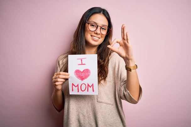 Giovane bella donna in possesso di carta con amore mamma messaggio che celebra la giornata delle madri facendo ok segno con le dita, simbolo eccellente
 - Foto, immagini