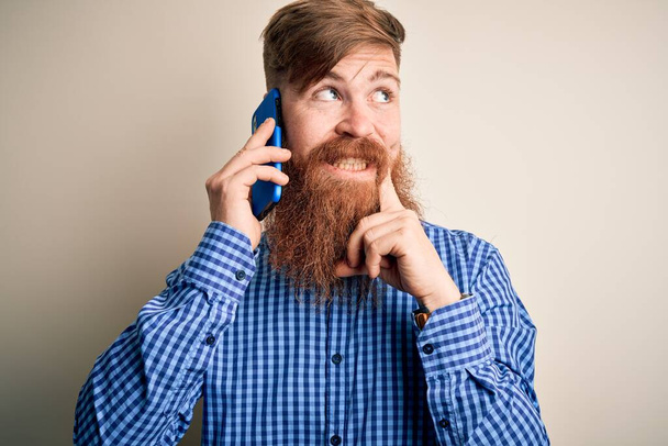 Rotschopf irischer Mann mit Bart unterhält sich auf dem Smartphone über isolierten Hintergrund, ernstes Gesicht denkt über Frage nach, sehr verwirrte Idee - Foto, Bild