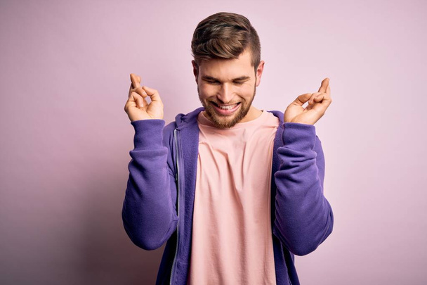 Jonge blonde man met baard en blauwe ogen dragen paarse sweater over roze achtergrond gebaren vinger gekruist glimlachend met hoop en ogen dicht. Geluk en bijgelovig concept. - Foto, afbeelding