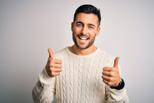 Fiatal jóképű férfi visel alkalmi pulóver felett áll elszigetelt fehér háttér siker jel csinál pozitív gesztus kézzel, hüvelykujj fel mosolyogva és boldogan. Vidám kifejezés és győztes gesztus. - Fotó, kép