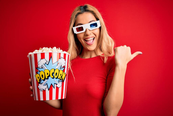 Jeune belle femme blonde regardant un film en utilisant des lunettes 3D manger des popcorns comme collation pointant et montrant le pouce sur le côté avec un visage heureux souriant
 - Photo, image