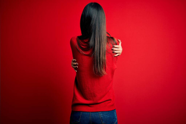 Jeune belle femme brune portant un pull décontracté sur fond rouge isolé S'embrasser heureux et positif à partir de l'arrière. Amour de soi et soin de soi
 - Photo, image