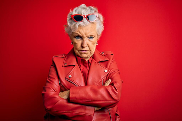 Senior mooie grijs-harige vrouw draagt casual rode jas en zonnebril sceptisch en nerveus, afkeurende uitdrukking op gezicht met gekruiste armen. Negatief persoon. - Foto, afbeelding