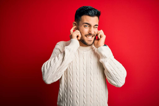 無声音楽のノイズにいらいらする表情で指で耳を覆う孤立した赤い背景の上に立つカジュアルな白いセーターを着た若いハンサムな男。ろう者の概念. - 写真・画像