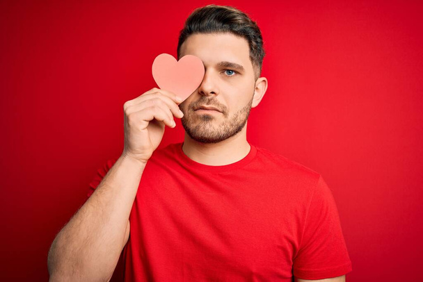 Mladý romantický muž s modrýma očima drží červené srdce papír ve tvaru nad červeným pozadím s sebevědomým výrazem na inteligentní tvář myšlení vážné - Fotografie, Obrázek