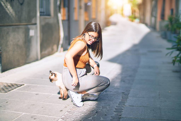 Mutlu ve kendinden emin gülümseyen genç ve güzel bir kadın. Yüzünde tebessümle dikilip kasaba caddesinde bir kediyi okşuyor. - Fotoğraf, Görsel