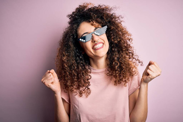 Young beautiful woman with curly hair and piercing wearing funny thug life sunglasses very happy and excited doing winner gesture with arms raised, smiling and screaming for success. Celebration concept. - Foto, Imagem
