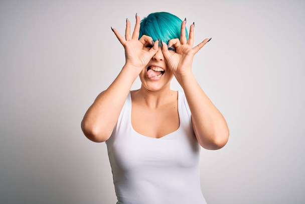 Νεαρή όμορφη γυναίκα με μπλε μαλλιά μόδας φορώντας casual t-shirt πάνω από λευκό φόντο κάνει ok χειρονομία, όπως κυάλια κολλήσει γλώσσα έξω, τα μάτια κοιτάζοντας μέσα από τα δάχτυλα. Τρελή έκφραση. - Φωτογραφία, εικόνα