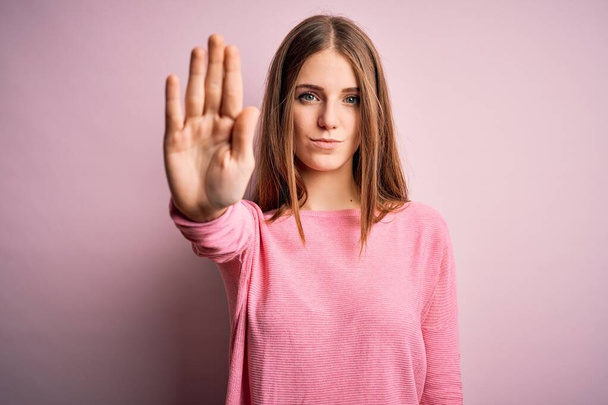孤立したピンクの背景にカジュアルなセーターを着た若い美しい赤毛の女性は、手の手のひらで歌うのをやめます。顔に負と深刻なジェスチャーで警告式. - 写真・画像
