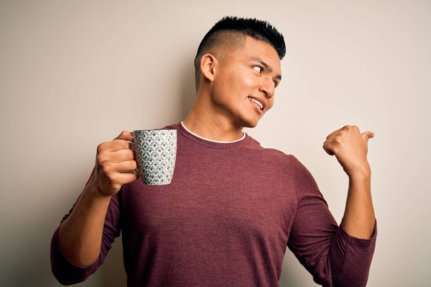 Nuori komea latinomies juo kupin kahvia yli eristetty valkoinen tausta osoittaa ja osoittaa peukalolla sivulle iloinen kasvot hymyillen
 - Valokuva, kuva