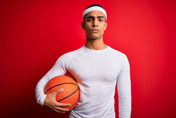 Young handsome african american sportsman holding basketball ball over red background with a confident expression on smart face thinking serious - Foto, Imagem