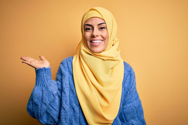 Junge schöne brünette muslimische Frau trägt arabischen Hijab über isoliertem gelben Hintergrund, lächelt fröhlich und zeigt mit der Handfläche in die Kamera. - Foto, Bild