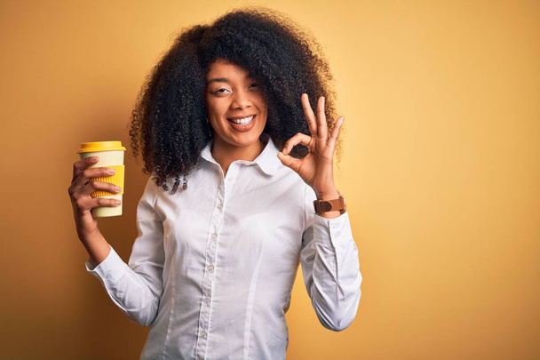 Νεαρή αφροαμερικανίδα επιχειρηματίας με αφρο μαλλιά πίνοντας καφέ από take away κύπελλο κάνει ok υπογράψει με τα δάχτυλα, εξαιρετικό σύμβολο - Φωτογραφία, εικόνα