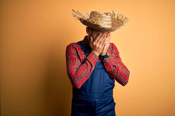 Nuori maaseudun viljelijä mies yllään ruokalappu yleistä ja maaseudun hattu keltainen tausta surullinen ilme peittää kasvot kädet itkiessään. Masennuksen käsite
. - Valokuva, kuva