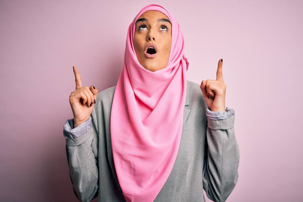 Young beautiful brunette businesswoman wearing pink muslim hijab and business jacket amazed and surprised looking up and pointing with fingers and raised arms. - Zdjęcie, obraz