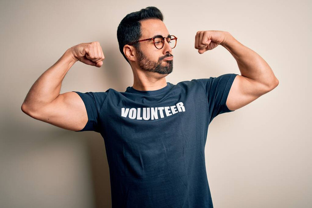 Homem bonito com barba vestindo camiseta com mensagem voluntária sobre fundo branco mostrando os músculos dos braços sorrindo orgulhoso. Conceito de aptidão
. - Foto, Imagem