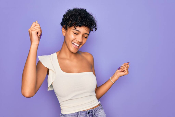 Νεαρή όμορφη Αφροαμερικανή Αφρικάνα που φοράει casual t-shirt πάνω από μωβ φόντο Χορεύει χαρούμενη και χαρούμενη, χαμογελαστή συγκινητική casual και με αυτοπεποίθηση ακούγοντας μουσική - Φωτογραφία, εικόνα