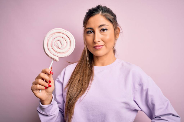 Młoda piękna brunetka kobieta jedząca słodkie cukierki na odizolowanym różowym tle z pewnym siebie wyrazem mądrej twarzy myśląc poważnie - Zdjęcie, obraz