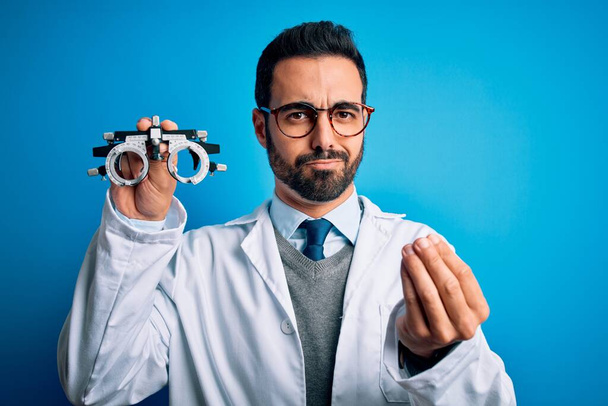 Junger gutaussehender optischer Mann mit Bart, der eine Optometriebrille über blauem Hintergrund hält, macht Geldgeste mit den Händen, verlangt Gehaltszahlung, Millionärsgeschäft - Foto, Bild