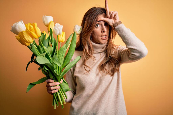 Young beautiful brunette woman holding bouquet of yellow tulips over isolated background making fun of people with fingers on forehead doing loser gesture mocking and insulting. - Photo, Image