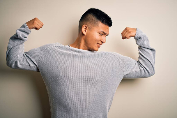 Ein junger gutaussehender Mann in lässigem Pullover steht vor isoliertem weißem Hintergrund und zeigt stolz lächelnde Armmuskeln. Fitnesskonzept. - Foto, Bild