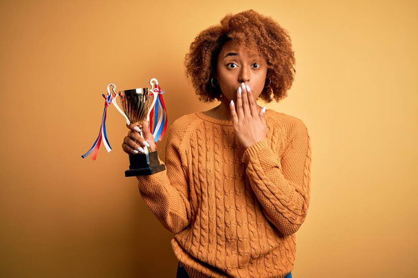 Young African American afro woman with curly hair holding winner champion trophy cover mouth with hand shocked with shame for mistake, expression of fear, scared in silence, secret concept - Photo, Image