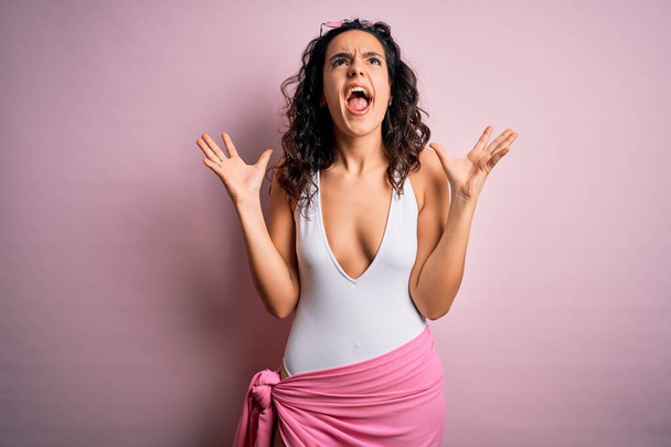Schöne Frau mit lockigem Haar im Urlaub trägt weißen Badeanzug über rosa Hintergrund verrückt und verrückt schreien und schreien mit aggressivem Ausdruck und erhobenen Armen. Frustrationskonzept. - Foto, Bild