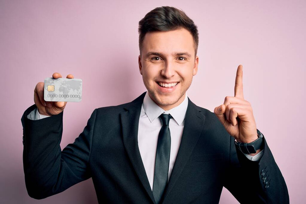 Jovem bonito homem de negócios caucasiano segurando cartão de crédito financeiro sobre fundo rosa surpreso com uma ideia ou pergunta apontando dedo com cara feliz, número um
 - Foto, Imagem