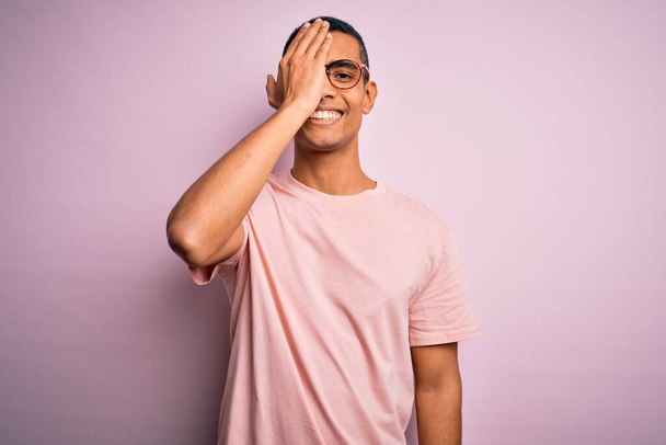 Bello uomo afroamericano che indossa casual t-shirt e occhiali su sfondo rosa che copre un occhio con mano, sorriso fiducioso sul viso ed emozione sorpresa
. - Foto, immagini