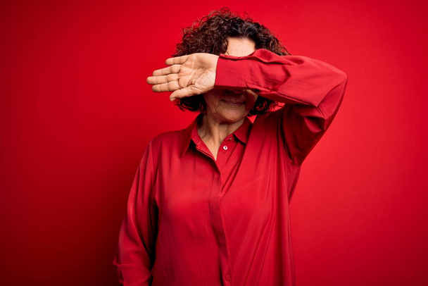 Middle age beautiful curly hair woman wearing casual shirt and glasses over red background covering eyes with arm, looking serious and sad. Sightless, hiding and rejection concept - Фото, изображение