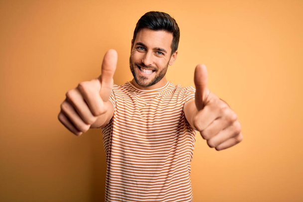 Young handsome man with beard wearing casual striped t-shirt over yellow background approving doing positive gesture with hand, thumbs up smiling and happy for success. Winner gesture. - Фото, изображение