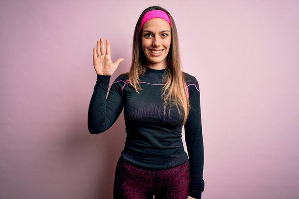 Młoda blondynka fitness kobieta noszenie sportowe ubrania treningowe na odosobnionym tle pokazując i wskazując palcem numer pięć uśmiechając się pewny siebie i szczęśliwy. - Zdjęcie, obraz